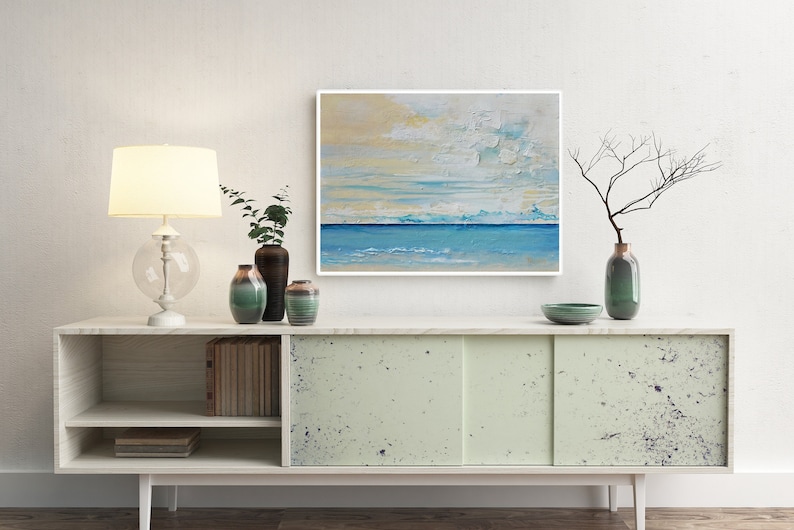 May include: A white cabinet with two sliding doors, a shelf, and a lower shelf. The cabinet is decorated with a light green paint with dark blue speckles. On top of the cabinet are various decorative items, including a lamp, a vase with a branch, and a bowl. A painting of a seascape with blue and yellow colours hangs on the wall behind the cabinet.