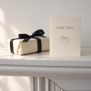 Puede incluir: Una tarjeta de felicitación de color crema con el texto FOR YOU y un dibujo lineal de una caja de regalo, colocada junto a un pequeño regalo rectangular envuelto en papel crema y atado con una cinta de satén negra, formando un conjunto.