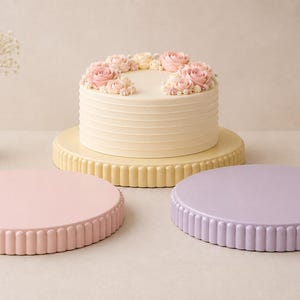 May include: A round, cream-colored cake with pink frosting flowers sits on a matching fluted cake stand. Two additional empty fluted cake stands in pastel pink and light purple are positioned in the foreground on a neutral surface.