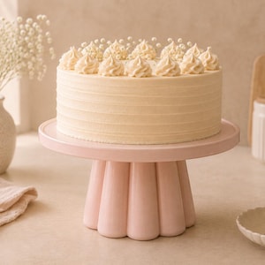 May include: A round cake with smooth cream-colored frosting and piped rosettes on top, displayed on a light pink ceramic cake stand with a fluted base. The cake features a textured side finish and small white decorative accents on the top surface.