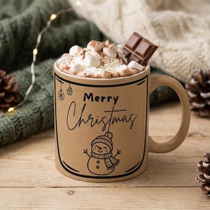 Puede incluir: Una taza de cerámica color canela con el texto negro Merry Christmas y un dibujo lineal de un muñeco de nieve. La taza está llena de chocolate caliente cubierto con malvaviscos blancos y un trozo de chocolate con leche en el borde.