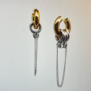 Puede incluir: Un par de pendientes de aro con una base circular dorada pulida. Cada pendiente está adornado con varios anillos plateados y una delicada cadena plateada colgante, creando un diseño de capas con una estética de metales mixtos.