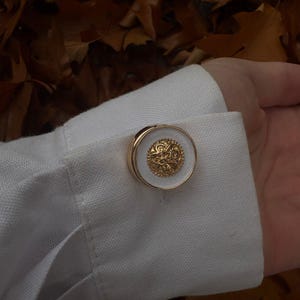 Op de afbeelding: Een ronde manchetknop met een centrale goudkleurige schijf met een ingewikkeld reliëfpatroon, omringd door een gladde witte rand en een goudkleurige metalen ring, bevestigd door de knoopsgaten van een witte stoffen overhemdmanchet.