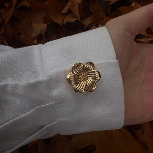 Pode incluir: Uma abotoadura de metal dourado em forma de flor de cinco pétalas, com uma superfície texturizada e canelada em cada pétala e um pequeno centro elevado. A abotoadura está presa através da casa do botão do punho de uma camisa branca.
