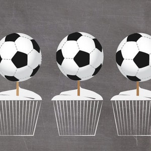 May include: Three cupcakes with white frosting and soccer ball toppers. The soccer balls are black and white, with a brown stick supporting each one. The cupcakes are in white paper liners, set against a gray background.