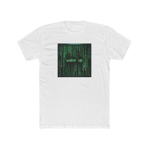 May include: A white short-sleeved crew neck t-shirt featuring a square graphic on the chest. The graphic displays a black computer screen filled with cascading green digital code and the text wake up in a pixelated green font centered.