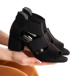 May include: A pair of black open-toe sandals featuring wide elastic straps across the top, a block heel, and a zipper closure at the back of the ankle. The footwear is constructed with a solid black finish and a peep-toe design.