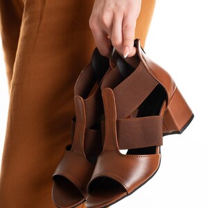 May include: A pair of brown open-toe sandals featuring a block heel, a T-strap design, and wide elastic side panels. The footwear is constructed with a smooth brown finish, a rounded toe opening, and a sturdy heel base for support and stability.