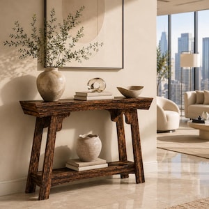 May include: A dark brown wooden console table with a rectangular top and a lower shelf. The table features angled legs and decorative brackets. It holds a large stone vase with green branches, a glass sculpture, a bowl, and stacked books.