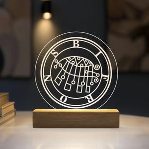 May include: A circular clear acrylic plaque featuring a white etched sigil of Bifrons with the letters B, I, F, R, O, N, S arranged around the perimeter. The plaque is mounted on a rectangular light-coloured wooden base that illuminates the design.