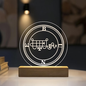 May include: A circular clear acrylic plaque featuring an etched sigil design with the letters E, B, O, and N positioned around the perimeter. The plaque is mounted on a rectangular light-coloured wooden base that illuminates the etched graphic.