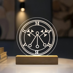 Könnte beinhalten: Ein kreisförmiges, klares Acryl-Lichtdisplay mit dem eingravierten Siegel von Marbas, wobei die Buchstaben M, A, R, B, A, S um den Rand angeordnet sind. Die Scheibe wird von unten durch einen rechteckigen hellen Holzsockel beleuchtet.