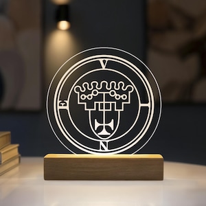 Könnte beinhalten: Eine kreisförmige, klare Acryl-Lichtplatte mit einem weiß geätzten Siegeldesign, das ein kronenartiges Symbol und die Buchstaben E, V, E, N am Rand zeigt, montiert auf einem rechteckigen, hellen Holzsockel, der das Design von unten beleuchtet.