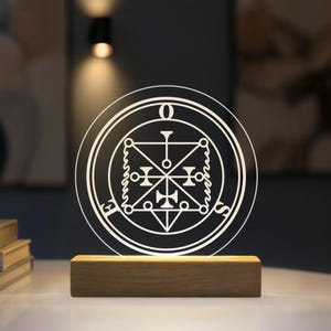Könnte beinhalten: Eine kreisförmige Acryl-Lichtplatte mit einem weiß geätzten Siegeldesign, bei dem die Buchstaben E, O und S um den Rand angeordnet sind. Die Platte ist auf einem rechteckigen Holzsockel montiert, der das Design von unten warm beleuchtet.