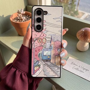 May include: A rectangular smartphone case featuring a printed illustration of a blue train travelling on tracks through a town with pink cherry blossom trees, a yellow speed limit sign, and a blue parking sign, with a triple camera cutout at the top.