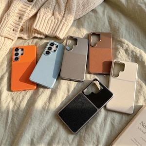 May include: A collection of six smartphone cases in orange, light blue, gray, brown, black, and cream. Each case features a smooth upper section and a textured, pebbled lower section, with a rectangular cutout for a multi-lens camera system.