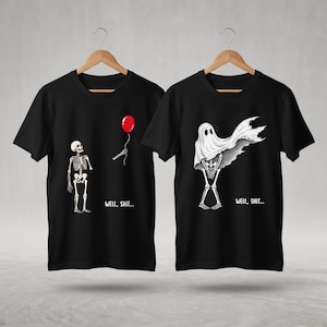May include: Two black t-shirts on wooden hangers. The left shirt features a skeleton holding a red balloon with the text "WELL, SHIT...". The right shirt shows a skeleton wearing a white ghost sheet with the text "WELL, SHIT..." printed below.