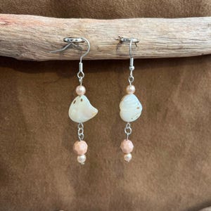 May include: A pair of drop earrings featuring silver-toned metal hooks, each suspending a small round peach-colored bead, an irregular white shell-like component, a larger faceted peach-colored bead, and a tiny matching peach-colored bead at the base.