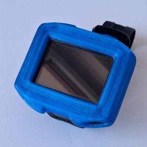 Peut inclure: Une coque de protection bleu vif installée sur un appareil électronique rectangulaire doté d'un écran noir. La coque présente une finition de surface texturée et superposée, et est fixée à une sangle noire visible en haut et en bas.