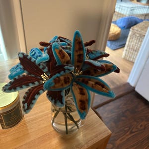 May include: A bouquet of handcrafted pipe cleaner flowers in a clear glass jar. The petals feature a brown and tan leopard-like spotted pattern with bright blue fuzzy edges. Each flower has a center with thin, light-colored stamens extending outward.