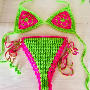 Può includere: Un set bikini all'uncinetto a due pezzi nei colori verde lime brillante e rosa acceso. Il top a triangolo presenta motivi floreali rosa, mentre lo slip è caratterizzato da un pannello centrale testurizzato verde lime con bordi smerlati rosa e laccetti laterali.