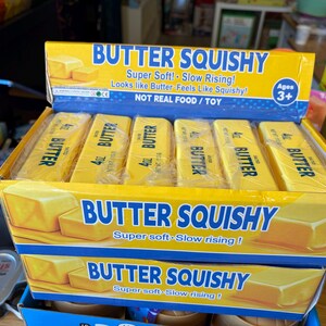 Puede incluir: Una caja expositora de cartón amarillo que contiene seis juguetes squishy con forma de mantequilla envueltos individualmente. Cada juguete está etiquetado como 4oz BUTTER. La caja presenta el texto BUTTER SQUISHY, Super Soft! Slow Rising! y NOT REAL FOOD / TOY.