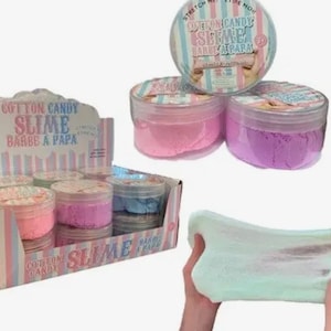 Puede incluir: Una caja expositora con varios botes de plástico transparente de slime Cotton Candy en rosa, morado y azul. Dos botes adicionales están apilados encima, y un slime de color verde claro, elástico y texturizado se muestra en primer plano.