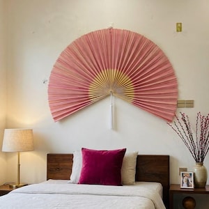 Peut inclure: Un grand éventail mural semi-circulaire composé de papier plissé rose avec un centre jaune clair. L'éventail est fixé au mur et comporte un pompon blanc suspendu au point central inférieur, créant un point focal décoratif au-dessus d'un lit.