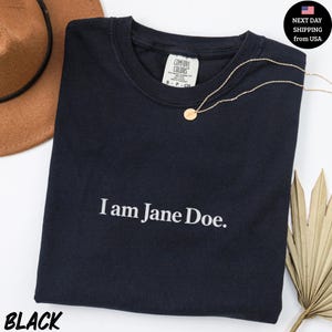 May include: A black short-sleeved t-shirt with the white text "I am Jane Doe." printed on the chest. The shirt features a Comfort Colors brand label at the inner neckline and is displayed alongside a brown hat and a gold necklace.