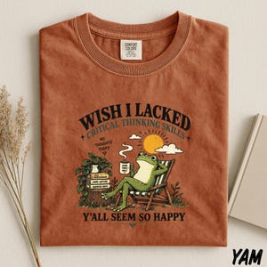 May include: A rust-colored t-shirt featuring a graphic of a green frog lounging in a beach chair, holding a mug labeled Good Vibes Only. The text reads Wish I Lacked Critical Thinking Skills, No Thoughts Today, and Y'all Seem So Happy.