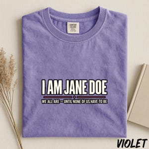 May include: A violet short-sleeved t-shirt with a crew neckline. The center of the shirt features the text I AM JANE DOE in white, followed by a horizontal line and the text WE ALL ARE - UNTIL NONE OF US HAVE TO BE in black.