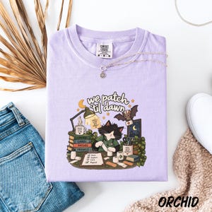 May include: A light purple t-shirt featuring a graphic of a cat dressed as a medical professional working at a desk. The text reads "We patch 'til dawn" with illustrations of books, a lamp, a bat, and medical supplies. The shirt has a Comfort Colors label.