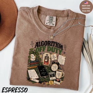May include: A brown crew neck t-shirt featuring a graphic print with the text ALGORITHM OFF DUTY. The design includes illustrations of a potted plant, a light switch, a stack of books, a smartphone, an alarm clock, a coffee mug, and notes.