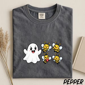 May include: A charcoal gray crew neck t-shirt with a graphic print featuring a white ghost and four yellow and black striped bees. One bee holds a small red heart. The shirt has a Comfort Colors brand label on the inside of the collar.