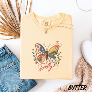 May include: A pale yellow t-shirt featuring an embroidered patchwork-style butterfly design with floral accents and the word Dolly in pink script. The shirt has a Comfort Colors label at the neck and is displayed with a gold chain necklace.