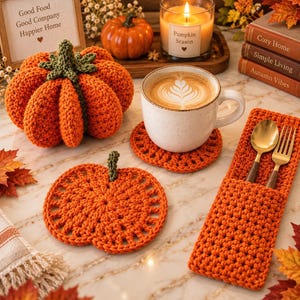 Op de afbeelding: Een set oranje gehaakte items waaronder een gevulde pompoen, twee ronde onderzetters en een rechthoekige bestekhouder met een gouden lepel en vork, gerangschikt met herfstdecoratie zoals boeken en een kaars.