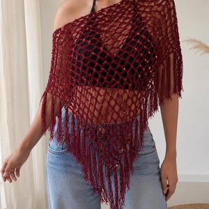 May include: A dark red crochet poncho featuring an open net pattern with integrated shimmering sequins and long fringe trim along the bottom edges. The garment is worn over a black halter top and light blue denim jeans, creating a layered look.