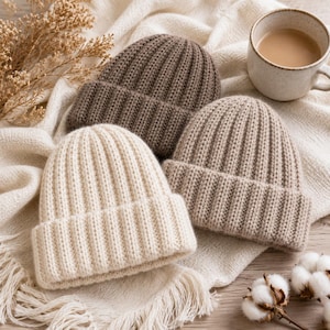 Pode incluir: Três gorros de malha canelada nas cores creme, bege claro e castanho escuro. Cada gorro apresenta uma dobra espessa e um padrão de malha vertical. Os gorros estão dispostos juntos, exibindo a sua textura macia e paleta neutra.