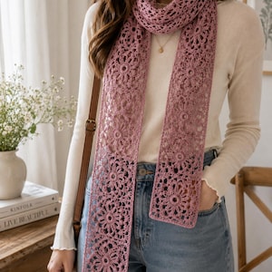 May include: A dusty rose crochet scarf featuring a repeating floral motif pattern. The scarf is constructed from interconnected circular lace elements, creating an openwork design that drapes vertically over a cream long-sleeve top and blue jeans.
