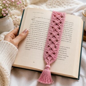 以下が含まれることがあります： 複雑なダイヤモンド型の格子模様と下部にタッセルが付いた、ダスティピンクのかぎ針編みのブックマーク。ブックマークは、ページにテキストが印刷された開いた本の中に配置されており、その細長い長方形の形状が示されています。