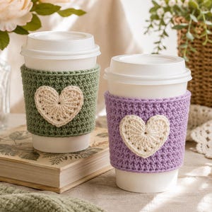 Puede incluir: Dos vasos de café desechables blancos con tapa, cada uno con una funda de ganchillo texturizada. Una funda es de color verde salvia y la otra es de color lavanda, ambas adornadas con un aplique de corazón de ganchillo color crema.
