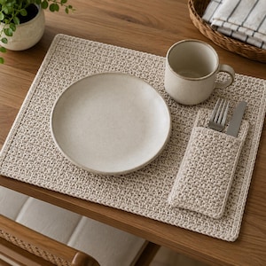 May include: A rectangular beige crochet placemat with a matching pocket for cutlery. The set includes a round off-white ceramic plate, a matching ceramic mug, a silver fork, and a silver knife. The items are arranged on a wooden surface.