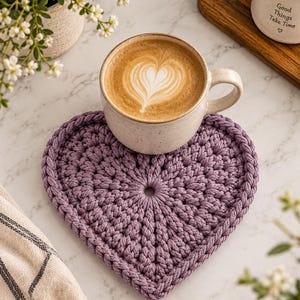 Könnte beinhalten: Ein herzförmiger Untersetzer aus dickem, violettem geflochtenem Kordelmaterial mit einer kreisförmigen Öffnung in der Mitte. Eine Keramiktasse mit einem Latte, der mit herzförmigem Milchschaum verziert ist, steht auf dem violetten Untersetzer.