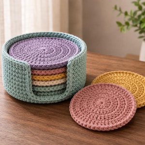 May include: A set of round, crocheted coasters in pastel shades of lavender, pink, yellow, and cream, stacked inside a matching light blue crocheted holder. Two additional coasters in pink and yellow are placed flat next to the holder.