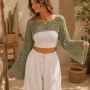 May include: A sage green crochet shrug featuring an open-knit pattern, a wide scoop neckline, and long, flared bell sleeves. The garment is worn over a plain white strapless tube top paired with white drawstring trousers.