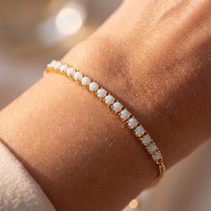 May include: A gold-toned tennis bracelet featuring a continuous row of small, round, iridescent white opal stones set in a metal chain. The bracelet is designed with a flexible link structure that wraps around the wrist, showcasing the stones.