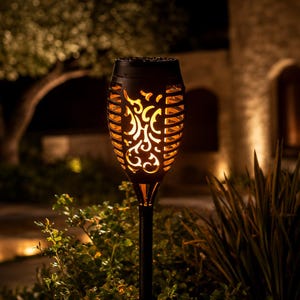 May include: A black solar-powered garden torch light featuring a cylindrical head with intricate cut-out patterns that emit a warm, flickering orange glow, mounted on a slender black stake designed for outdoor ground installation.