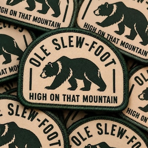 May include: A rectangular patch with rounded top corners featuring a dark green bear silhouette on a tan background. The text OLE SLEW-FOOT is arched above the bear, and the text HIGH ON THAT MOUNTAIN is printed in dark green below the bear.
