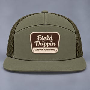 May include: An olive green trucker hat with a mesh back and a flat brim. A rectangular brown patch is centered on the front panel, featuring the white text Field Trippin and the smaller text OUTDOOR PLAYGROUND below it.