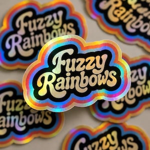 May include: A cloud-shaped sticker with a holographic finish featuring the text Fuzzy Rainbows in a bold, rounded font. The design includes a black center with the text surrounded by concentric bands of red, orange, yellow, green, and blue.